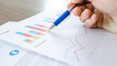 A close-up of a hand with a pen analyzing data on colorful bar and line charts on paper.