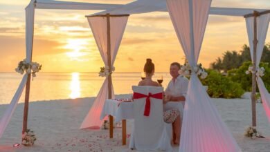 A couple enjoys a romantic dinner on a beach under a canopy at sunset, surrounded by a serene ocean view.