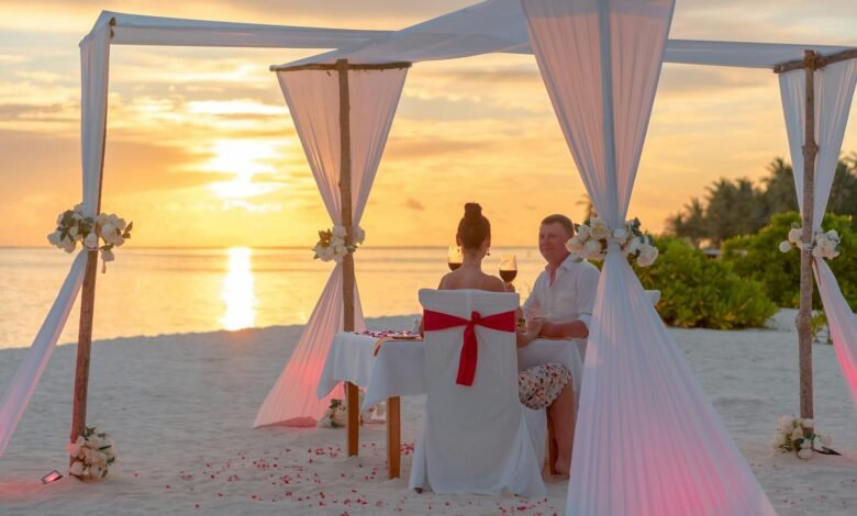 A couple enjoys a romantic dinner on a beach under a canopy at sunset, surrounded by a serene ocean view.